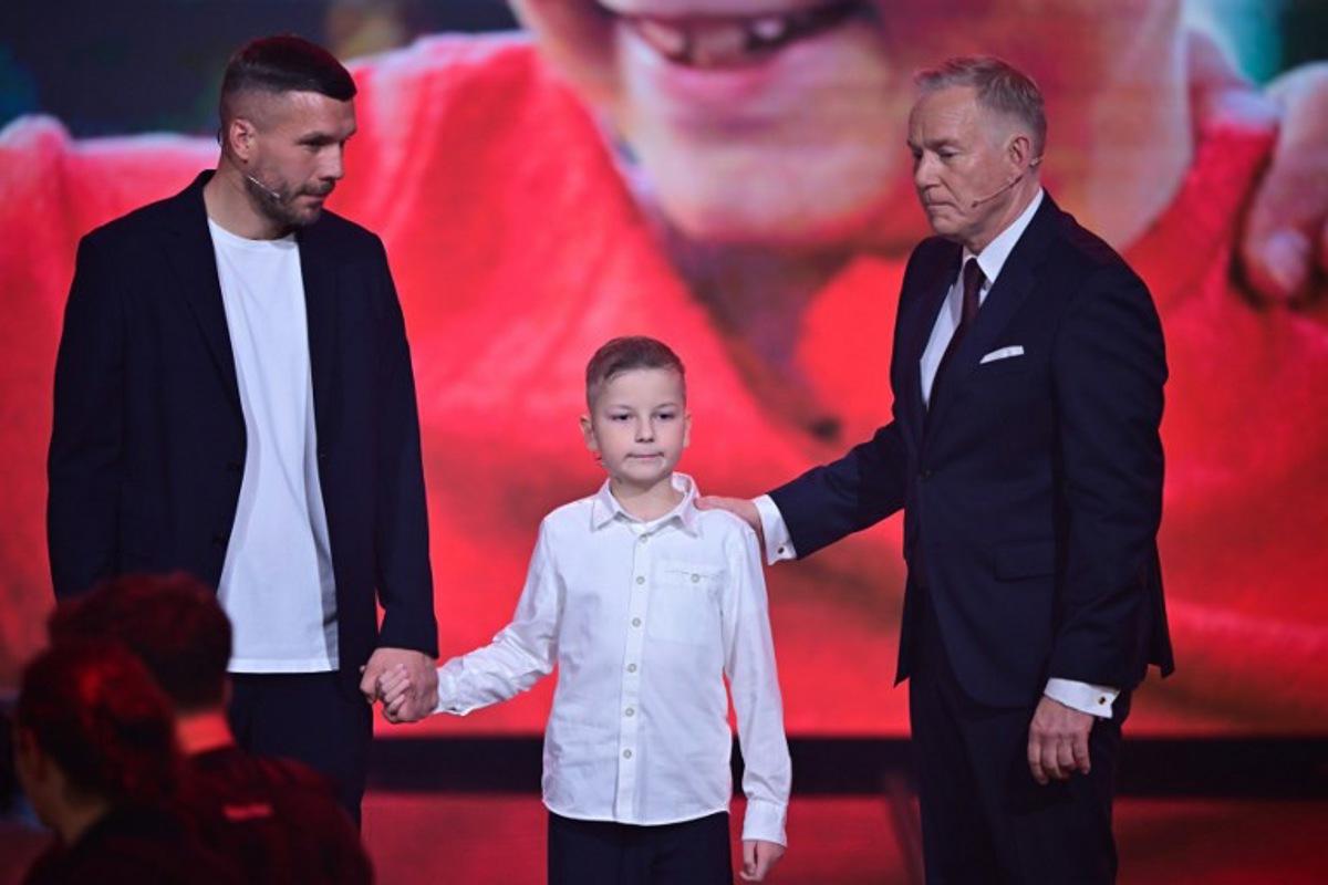 German TV host Johannes B. Kerner (R), cancer survivor Collin (C) and former German football player Lukas Podolski stand on stage during the charity gala "Ein Herz Für Kinder" (A heart for children) in Berlin, on December 6, 2025. Sebastian Gollnow / POOL / AFP